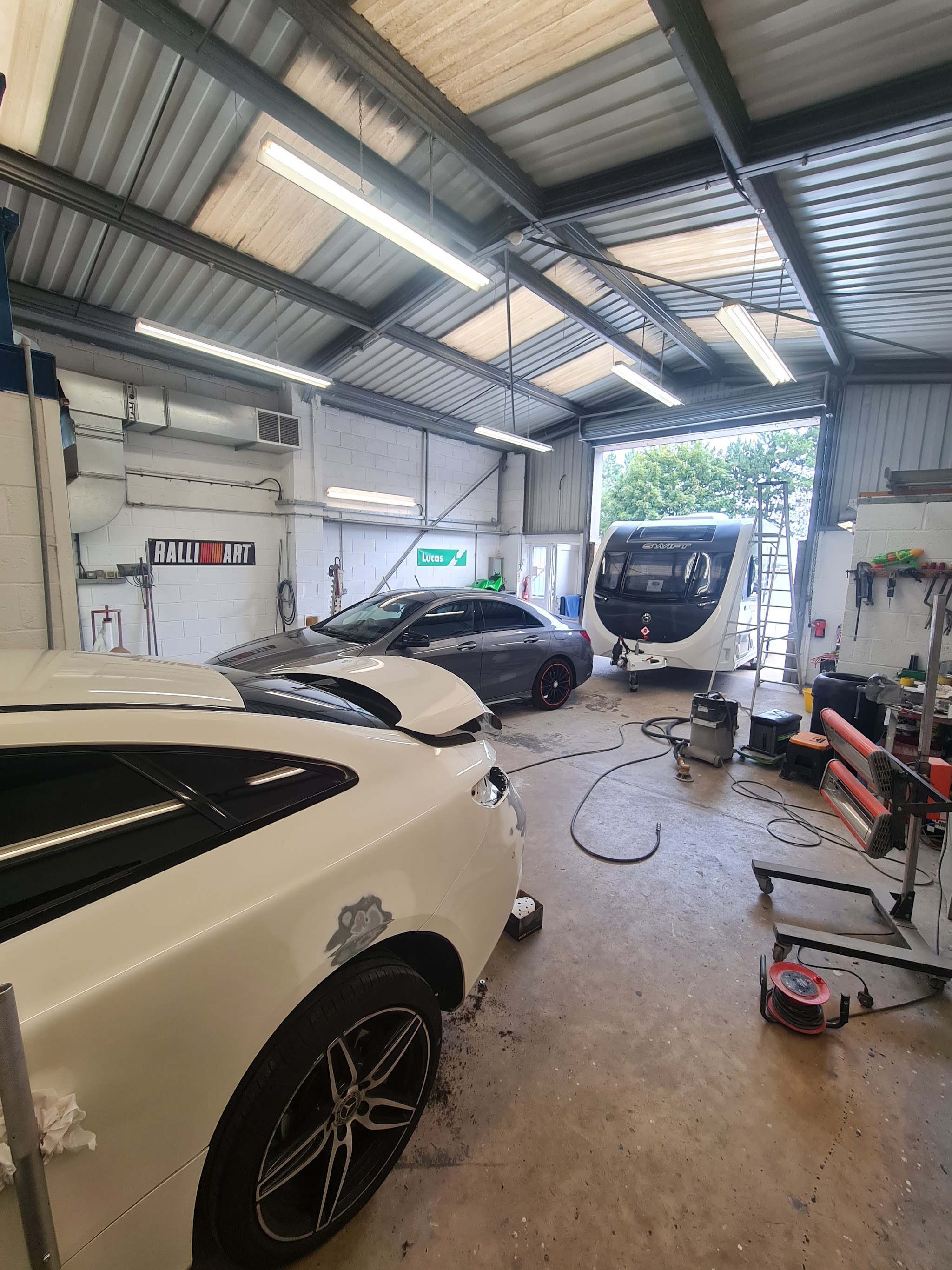 Inside the workshop: a white car being prepared, a grey Mercedes, and a Swift caravan by the open roller door